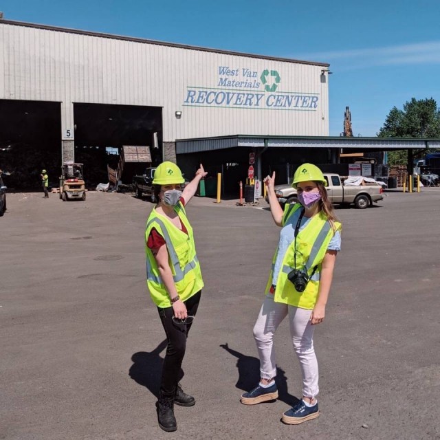 Marissa Porcellini and Shannon Hunter point at the West Van Materials Recovery Center while standing in front of the building on a blue sky day. Cars and machinery are in motion in the background depositing materials.